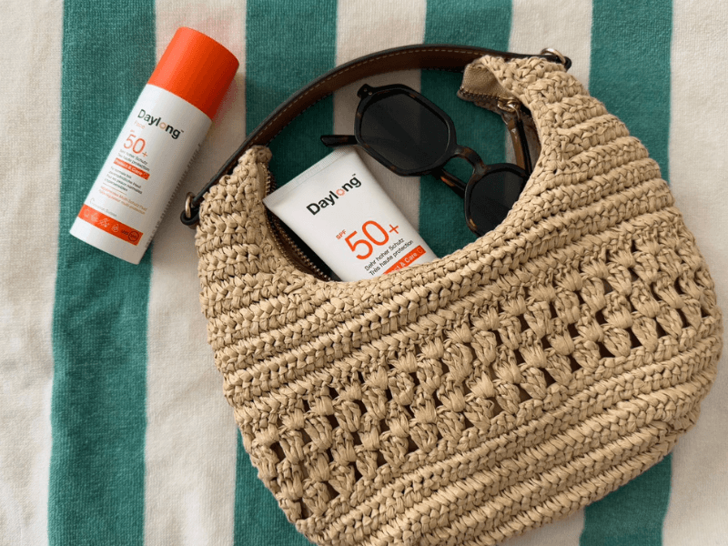 Sommeressentials: Sonnenschutz von Daylong Protect&Care in Basttasche auf grün-weiss-gestreiftem Handtuch.